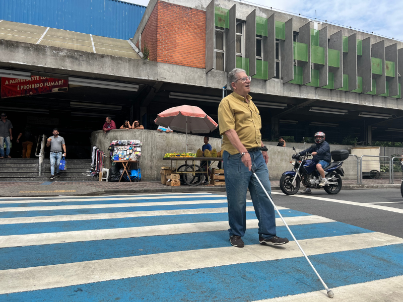 Pessoa com deficiência visual atravessando a rua para chegar à Estação Prefeito Celso Daniel